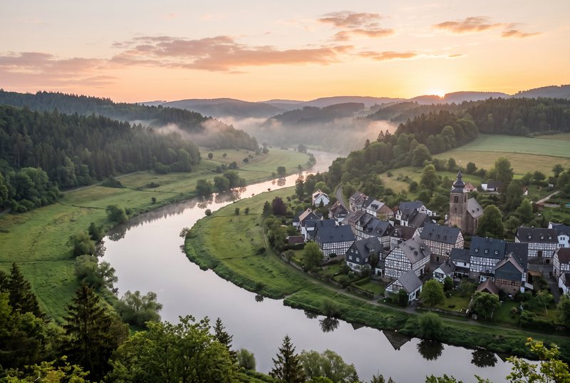 Fluss mit Fachwerkstädtchen in einem grünen Tal bei Sonnenaufgang